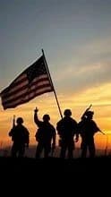 Four soldiers silhouetted at sunset, one holding the American flag
